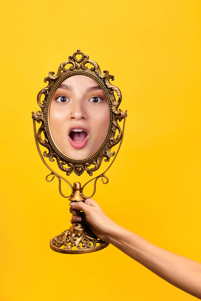 Wow. Female hand holding mirror with reflection of shouting girls face isolated over yellow background. Concept of vintage, retro style, beauty, art, creativity and ad. Human emotions, facial - Photo, image