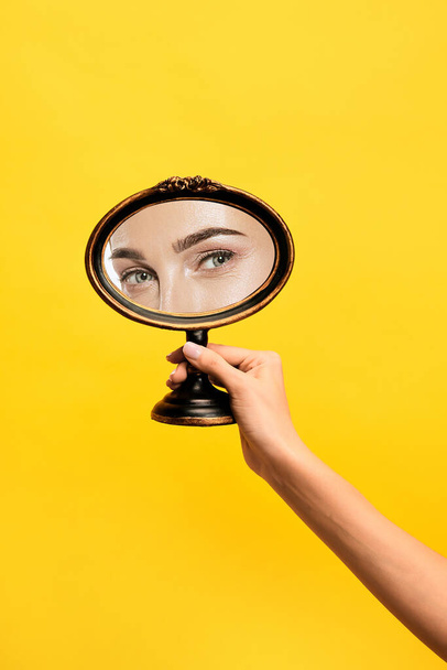 Happy look. Female hand holding mirror with reflection of girls eyes isolated over yellow background. Concept of vintage, retro style, beauty, art, creativity and ad. Human emotions, facial - Фото, изображение