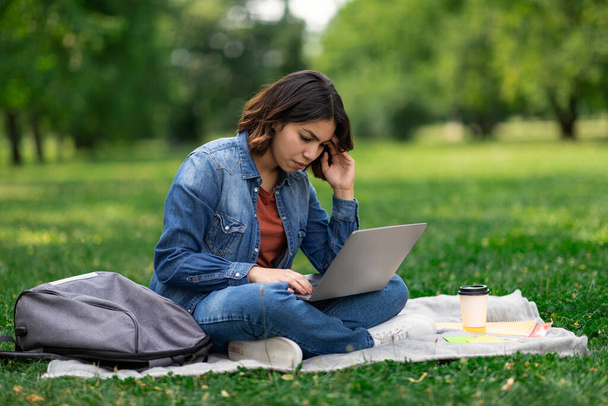 Upset Arab Female Student Using Laptop While Sitting On Plaid In Park, Young Middle Eastern Woman Study With Computer Outdoors At Campus, Having Problems With Distance Learning, Copy Space - 写真・画像