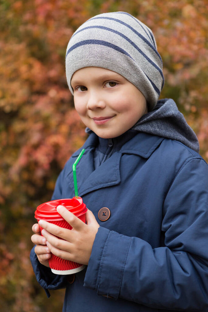 ritratto di un ragazzo con un bicchiere di cartone rosso, passeggiare nel parco autunnale o nella foresta, tempo freddo, bevanda calda in un bicchiere, concetto - Foto, immagini