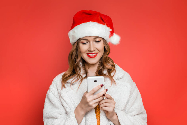 Mujer caucásica feliz en sombrero de Navidad y abrigo de piel sintética blanca sosteniendo el teléfono móvil en sus manos, posando en el estudio y mensajes de texto aislados sobre fondo rojo - Foto, Imagen