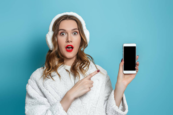 Shocked surprised young woman in white faux fur coat points finger at mobile phone screen isolated on blue studio background. Mo?kup for design, text, empty space - Foto, immagini