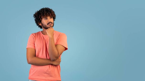 Pensive atraktivní kudrnatý mladý indián v růžovém tričku při pohledu na kopírovací prostor pro text nebo reklama na modrém pozadí, dotýkání brady, studiový záběr, panorama s prázdným prostorem - Fotografie, Obrázek