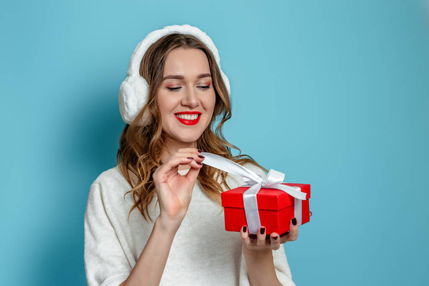 Online gift order 2021. Close up portrait of young smiling caucasian woman holding and opening red gift box isolated on blue studio background - Photo, Image