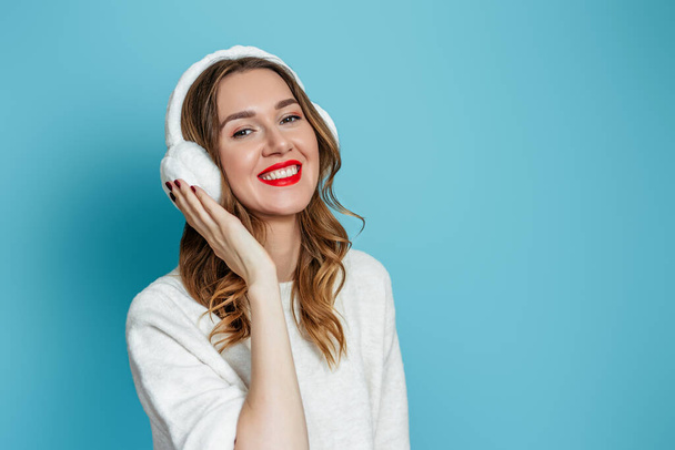Happy young caucasian woman in fur headphones and white sweater isolated on blue studio background. Copy space - Photo, Image