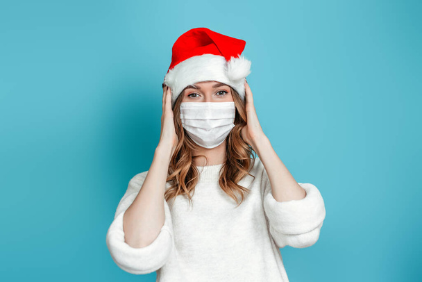 Brunette woman wearing protective medical face mask and christmas holiday hat holding her head isolated on blue studio background. 2021 new year concept - Photo, image