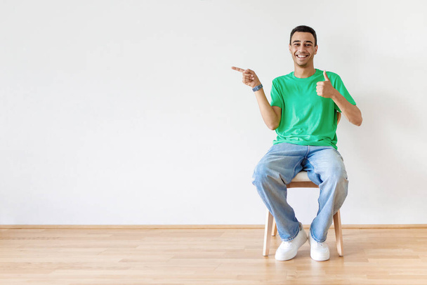 Hombre árabe feliz gestos pulgar hacia arriba y señalar el dedo a un lado en el espacio libre, sentado en la silla sobre el fondo de la pared de luz. Me gusta esta oferta y concepto de promoción - Foto, imagen
