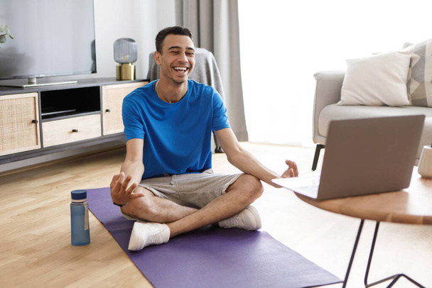Calme positif homme arabe en vêtements de sport assis dans la pose de lotus, ayant cours de yoga en ligne, méditant près d'un ordinateur portable à la maison. Jeune gars suivant tutoriel en ligne, faire de la forme physique domestique - Photo, image