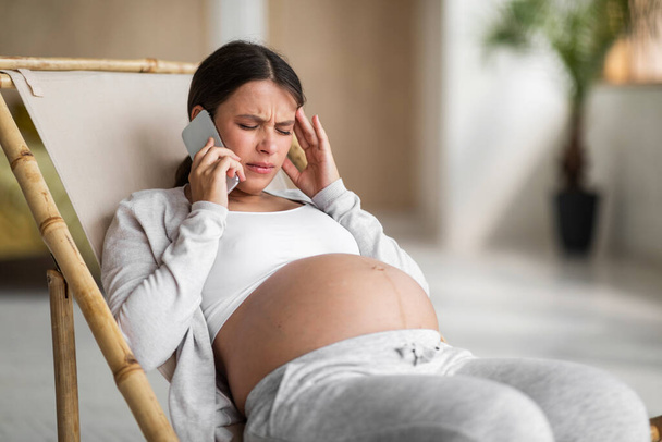 Nuori raskaana oleva nainen kärsii päänsärystä puhuessaan matkapuhelimella kotona, järkyttynyt odottava nainen istuu tuolissa ja kukkii, tunne huonovointisuus, akuutti migreeni, lähikuva laukaus vapaalla tilalla - Valokuva, kuva