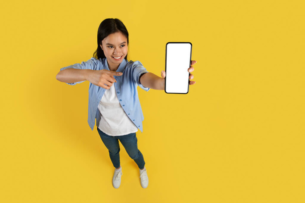 Mosolygó vicces fiatal kínai lány diák alkalmi mutogató ujját okostelefon üres képernyőn, elszigetelt sárga háttér, stúdió. Hirdetés és ajánlat, hatalmas eladó, alkalmazás az online vásárlás és a győzelem - Fotó, kép