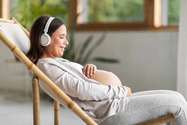 美しい妊娠中の女性が椅子でリラックスし、ワイヤレスヘッドフォンで音楽を聞きます,笑顔の側のビューショット期待される女性は好きな曲を楽しんで、腹を包含します,スペースをコピー - 写真・画像