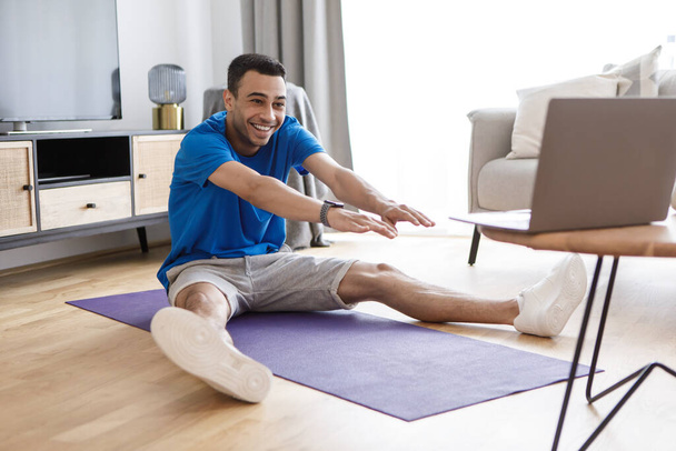 Sessão de treino online. Homem árabe feliz alongamento pernas músculos e quads enquanto assiste tutorial on-line no pc, sentado no tapete, exercitando-se na sala de estar interior - Foto, Imagem