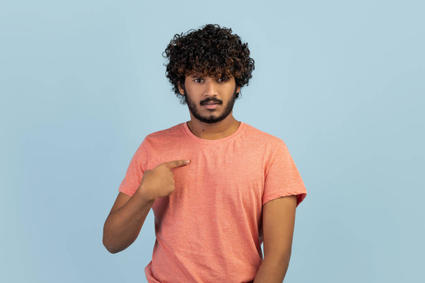 Are you talking about me. Questioned not sure cute handsome curly bearded indian man, looking surprised pointing himself, making sure he heard right, being chosen, posing over blue background - Foto, imagen