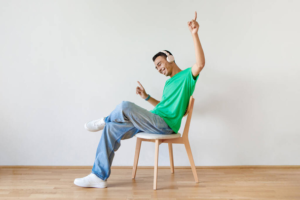 Cheery guy in headphones listening to music and dancing while sitting on chair against white studio wall, banner design with free space. Joyful man moving to favorite song - Photo, Image