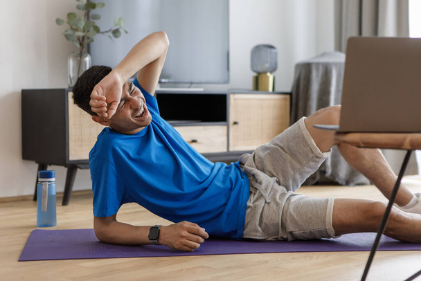 Fitness-Burnout. Müder arabischer Mann fühlt sich schlecht nach Online-Workout, Training mit Laptop, zu Hause auf Matte liegend. Sporteinsteiger beim Turnen in der Halle - Foto, Bild