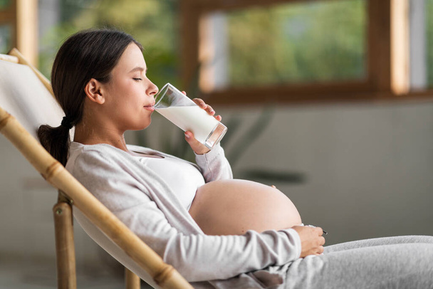 Junge schöne schwangere Frau ruht sich im Stuhl aus und trinkt Milch aus dem Glas, Seitenansicht Aufnahme der attraktiven werdenden Millennial Frau, die zu Hause gesundes Kalziumgetränk genießt, Kopierraum - Foto, Bild