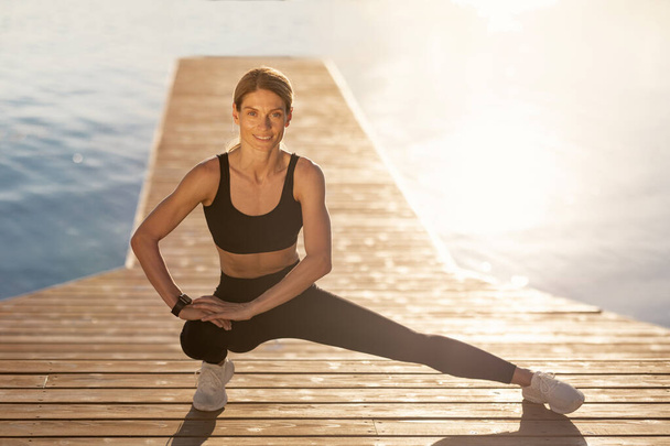 Middle Aged Athletic Woman Exercising On Wooden Pier Outdoors, Smiling Sporty Female In Activewear Making Side Lunges Exercise While Training Outside, Enjoying Fitness Lifestyle, Copy Space - Photo, Image