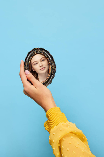 Young redheaded girl holding vintage style mirror in her hands and looking at her reflection. Concept of beauty, emotions, art. Colorful minimalism. Blue and yellow - Fotografie, Obrázek