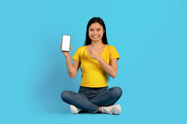 Sarı tişörtlü, gülümseyen genç Koreli kadın lotus pozisyonunda oturuyor, akıllı telefonu boş ekranla işaret ediyor, mavi arka planda, stüdyoda izole edilmiş. Çalışma, reklam ve iş için yeni uygulama - Fotoğraf, Görsel