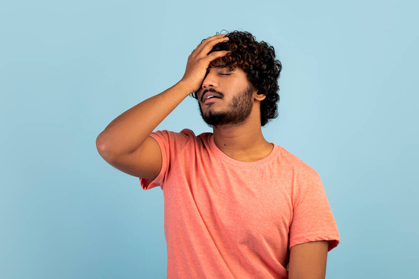 Sad curly millennial eastern guy in casual outfit touching his head, eyes closed, unhappy exhausted young indian man having troubles, posing over blue studio background, copy space - Photo, Image