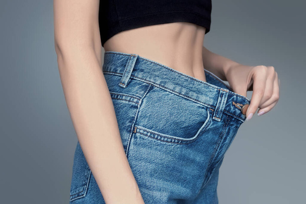 Thin woman body in big jeans isolated on a gray background. Concept of weight loss. Girl holds jeans in her hand and shows a thin waist. Woman in oversize jeans, diet concept - Foto, Bild