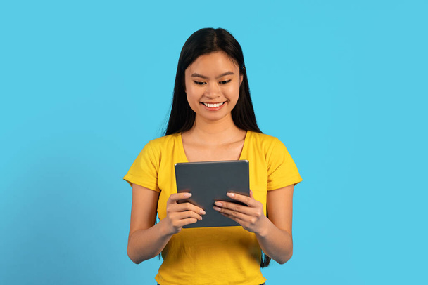 Ευτυχισμένη έξυπνη πολυάσχολη νεαρή Κινέζα μαθήτρια με κίτρινο μπλουζάκι δακτυλογραφώντας σε tablet, απομονωμένη σε μπλε φόντο, στούντιο. Εργασία εξ αποστάσεως, αγγελία και προσφορά, εφαρμογή για το blog και την τεχνολογία για μελέτη λόγω covid-19 - Φωτογραφία, εικόνα