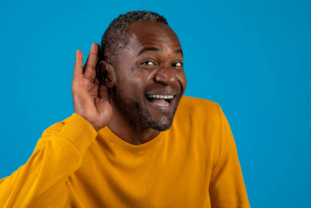 Ενθουσιασμένος μεσήλικας μαύρος άντρας με κίτρινο χέρι δίπλα στο αυτί και χαμογελώντας, ακούγοντας ειδήσεις, κουτσομπολιά ή φήμες, ποζάροντας πάνω από το μπλε φόντο του στούντιο, κοντινό πλάνο, αντιγραφή χώρου - Φωτογραφία, εικόνα