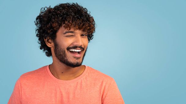Veselý stylový pohledný vousatý mladík s kudrnatými vlasy pózující na modrém pozadí studia, mrkající a usmívající se na kameru, panorama s kopírovacím prostorem, detailní záběr. Millennials životní styl koncept - Fotografie, Obrázek
