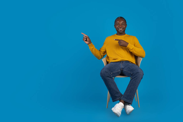 Relaxed happy handsome mature black man in casual outfit sitting on chair over blue studio background, showing copy space for advertisement and smiling, gesturing, panorama - Photo, Image