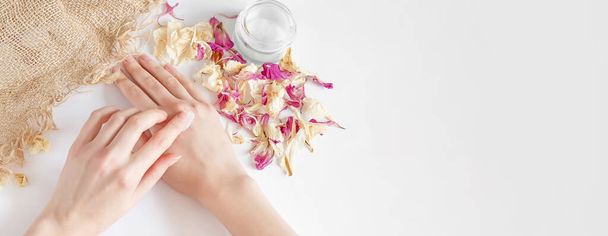 web banner. woman applies moisturizer to her hands on a white background with dry flower petals and a piece of burlap, top view. Ecological, natural fragrance-free cream, natural hand care oil. - Photo, Image