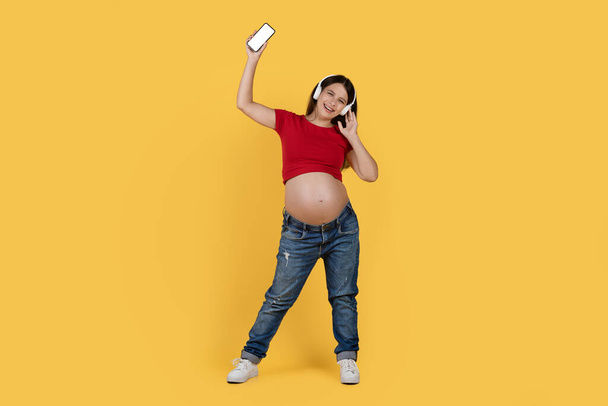 Happy young pregnant woman in headphones listening music on smartphone and dancing over yellow studio background, cheerful expectant female having fun, enjoying favorite playlist, copy space - Foto, Bild