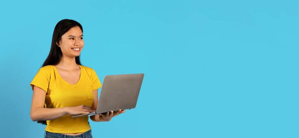 Улыбающаяся умная китаянка в желтой футболке учится за ноутбуком, изолирована на синем фоне, панорама, студия. Технологии для работы, рекламы и предложения, цифровые кочевники и фрилансеры - Фото, изображение