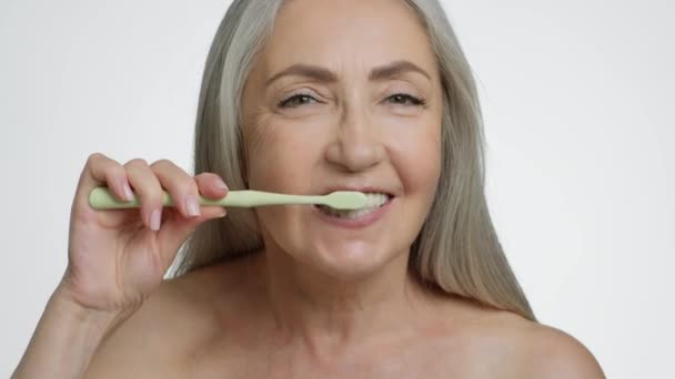 Tandheelkundige zorg op hoge leeftijd. close-up portret van positieve oudere vrouw poetsen haar tanden met natuurlijke borstel, kijken naar camera, witte studio achtergrond, slow motion - Video