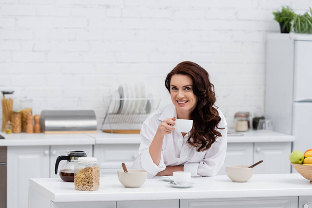 Femme souriante en chemise tenant une tasse de café près des céréales à la maison  - Photo, image