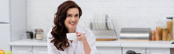 Smiling brunette woman in shirt holding cup of coffee in kitchen, banner  - Foto, imagen
