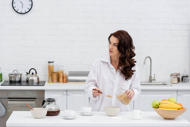Brunette woman in shirt holding cereal rings near coffee and fruits in kitchen  - Photo, Image