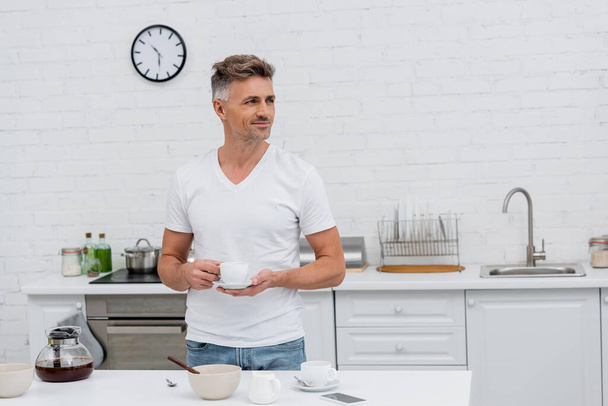 Lächelnder Mann im T-Shirt hält Tasse Kaffee neben Smartphone in der Küche  - Foto, Bild