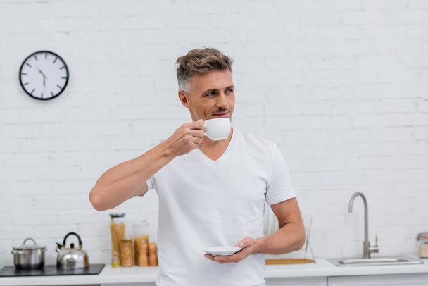 Man in t-shirt holding cup of coffee in kitchen in morning  - Fotó, kép