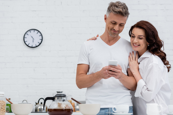 Positive woman hugging husband with cellphone near coffee and breakfast in kitchen  - Фото, изображение