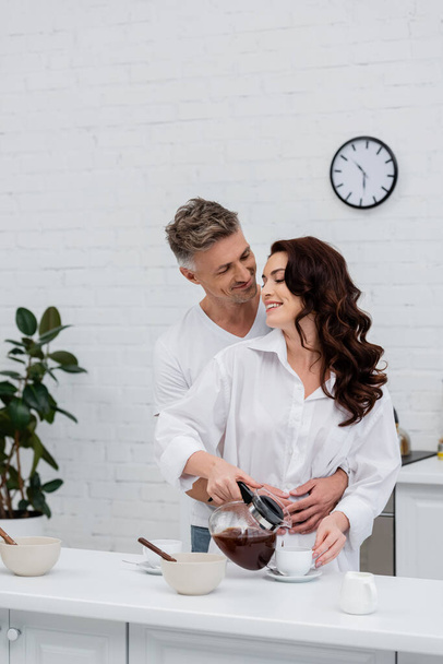 Smiling man hugging brunette wife in shirt pouring coffee in kitchen  - Zdjęcie, obraz