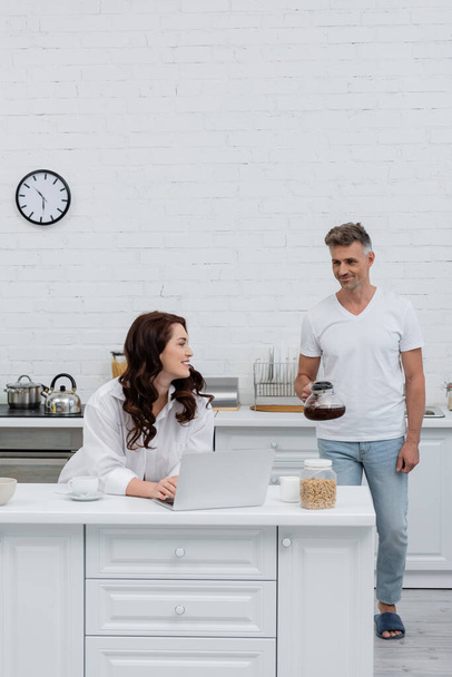 Smiling woman using laptop near husband with coffee pot and cereal rings in kitchen  - Photo, Image