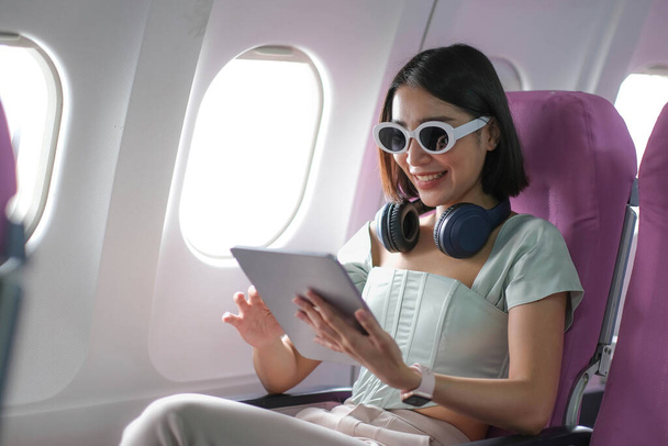 Reisetourismus mit moderner Technik und Flugkonzept, Frau sitzt im Flugzeug mit modernem digitalen Gerät und sucht Lieblingsmusik-Playlist in Anwendung für Hörer - Foto, Bild