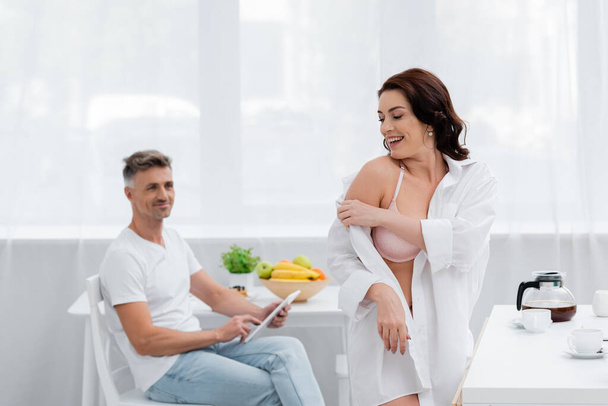 Mujer sonriente en sujetador quitándose la camisa cerca del café y marido borroso con tableta digital en la cocina  - Foto, imagen