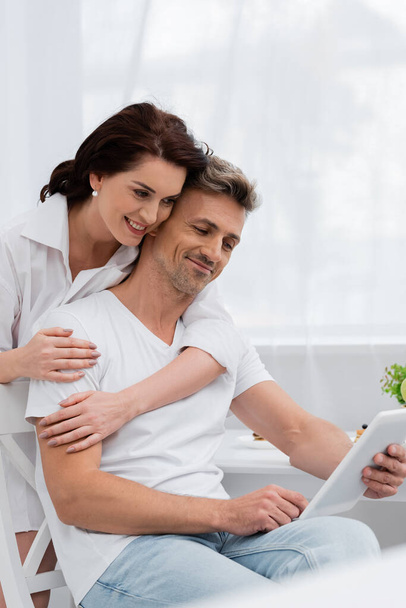 Mosolygós nő ingben átöleli férj segítségével digitális tabletta a konyhában  - Fotó, kép