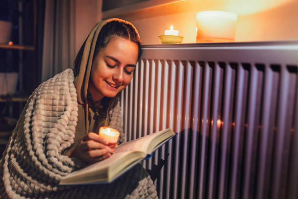 Teini-ikäinen tyttö istuu peiton alla lähellä lämmityspatteria kynttilöillä ja lukea kirjaa.Kasvavat kustannukset yksityisissä kotitalouksissa kaasulaskun takia inflaatio ja sota, energiakriisi - Valokuva, kuva