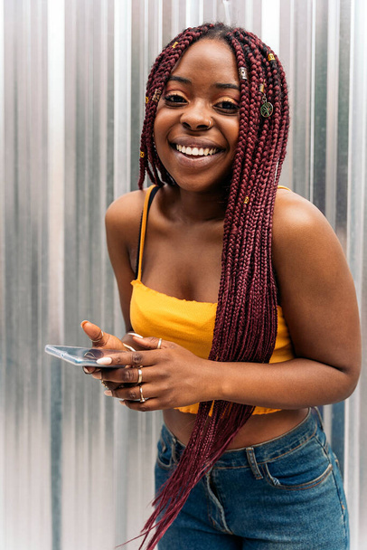 Cheerful young african woman with braids looking at camera and holding her smart-phone. - Photo, image
