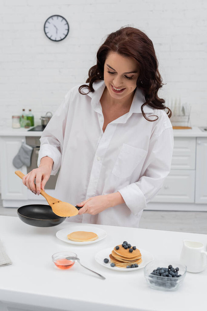 Smiling woman in shirt cooking pancakes near blueberries in kitchen  - Foto, Bild