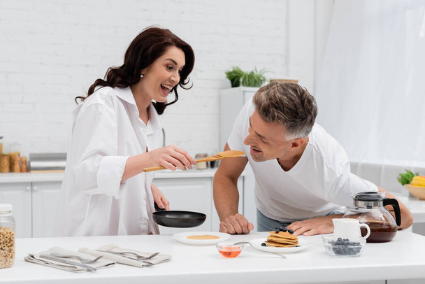 Smiling woman feeding husband with pancake near breakfast in kitchen  - 写真・画像