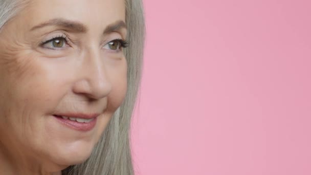 Aging and natural beauty. Close up portrait of beautiful grey haired senior lady with wrinkles around eyes looking aside and smiling, pink studio background, slow motion, empty space - Footage, Video