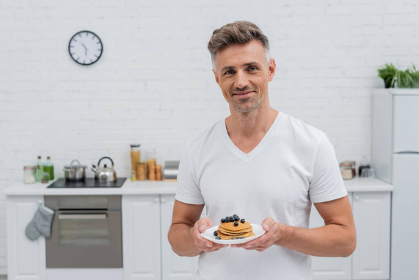 Smiling man looking at camera and holding pancakes with blueberries in kitchen  - Φωτογραφία, εικόνα